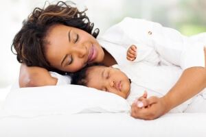 Mother dressed in white, cuddling happily with their baby on a white bed.